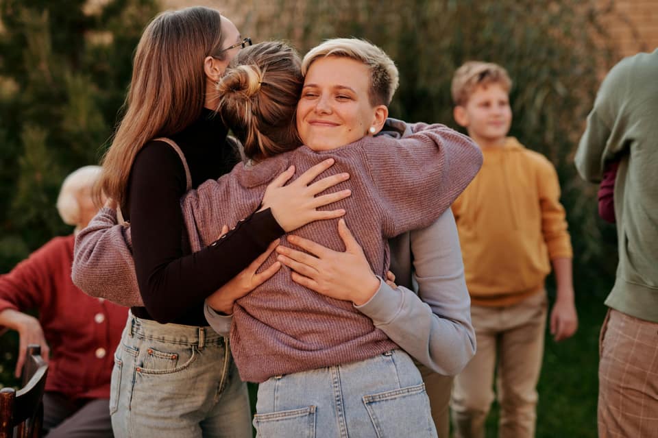 Four people hugging outdoors, with others blurred in the background