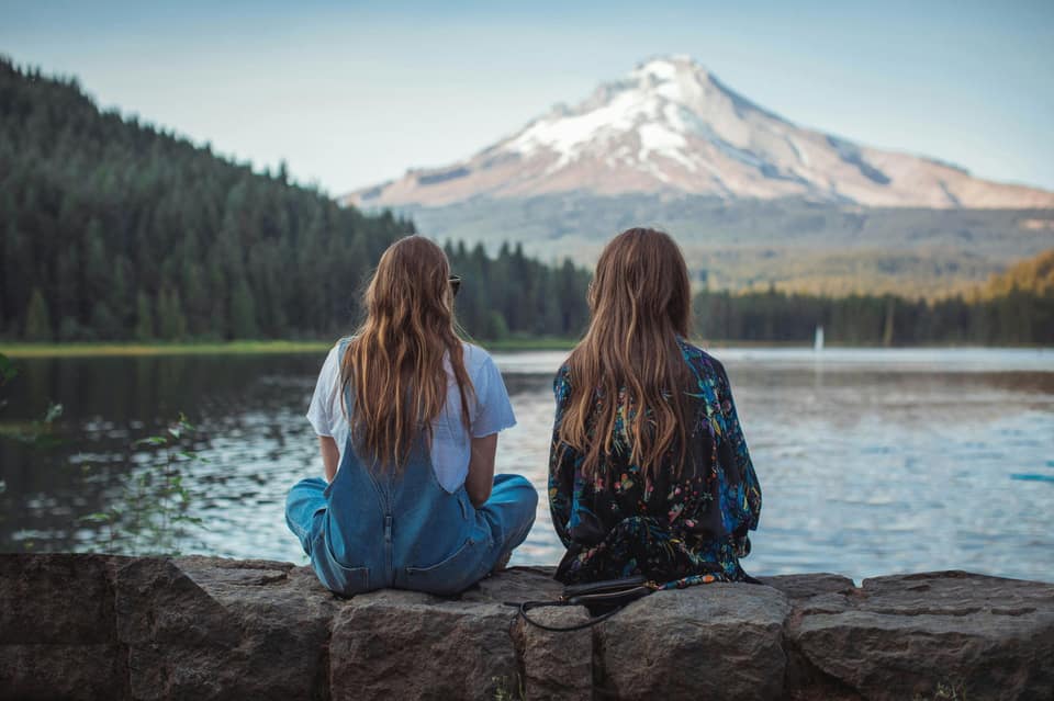 Two women sitting on rocks by a lake, looking toward a snow-capped mountain
